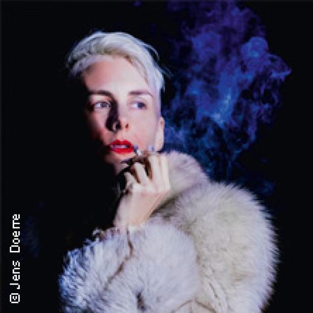 Eine Person mit weißblondem Haar, rotem Lippenstift und Pelzmantel raucht eine Zigarette vor dunklem Hintergrund.