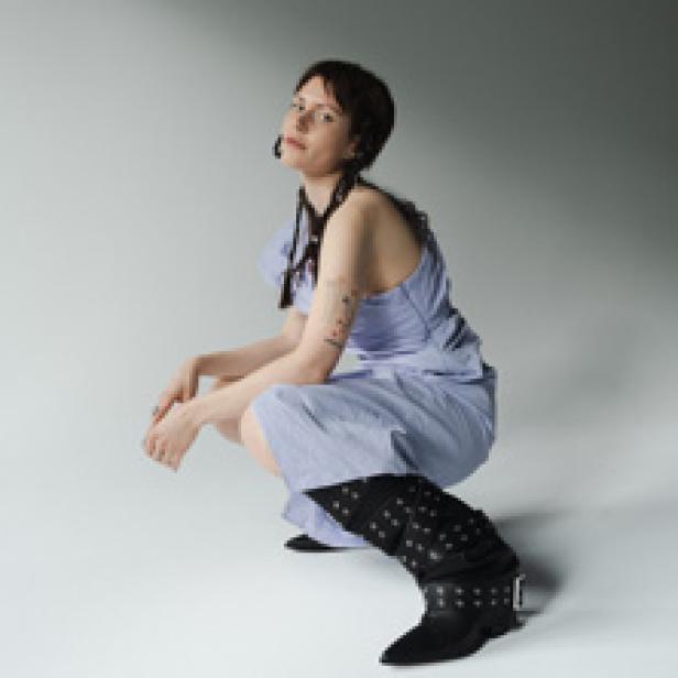 Eine Frau in einem hellblauen Kleid und Stiefeln posiert vor einem grauen Hintergrund.