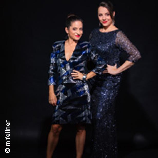 Zwei Frauen posieren in glitzernden Kleidern vor einem schwarzen Hintergrund.