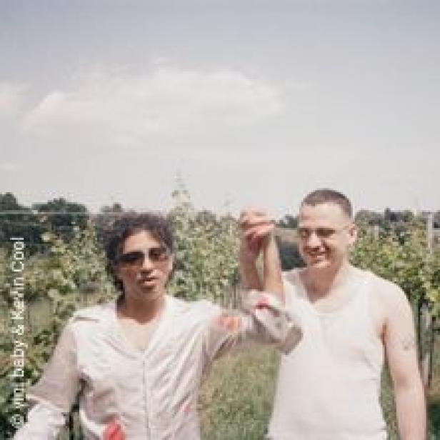 Zwei Männer stehen vor einem Weinberg und halten Händchen.