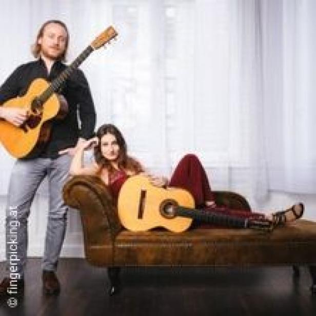Ein Mann steht mit einer Gitarre, während eine Frau mit einer Gitarre auf einer Couch liegt.