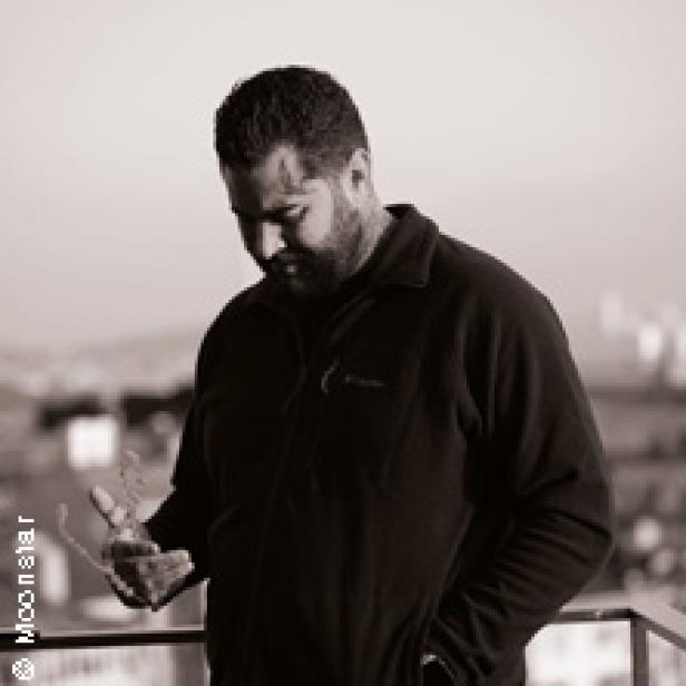 Ein Mann mit Bart steht auf einem Balkon und blickt nach unten.