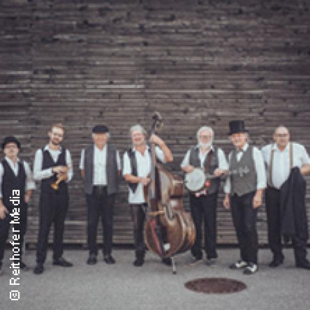 Eine Band mit sieben Mitgliedern posiert mit ihren Instrumenten vor einer Holzwand.