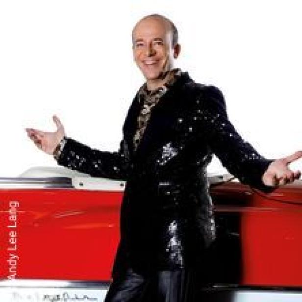 Ein Mann in einem glitzernden Sakko steht vor einem roten Auto und lächelt.