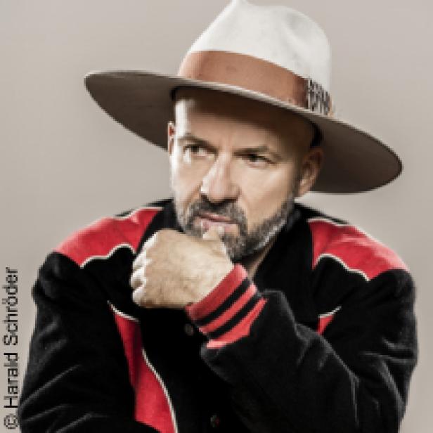 Ein Mann mit Hut und Jacke posiert vor einem hellen Hintergrund.