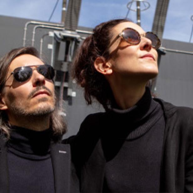 Ein Mann und eine Frau mit Sonnenbrillen stehen vor einem Gebäude und schauen nach oben.