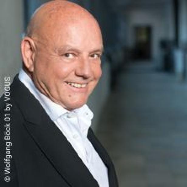 Ein lächelnder, kahlköpfiger Mann in einem schwarzen Anzug steht vor einem dunklen Hintergrund.