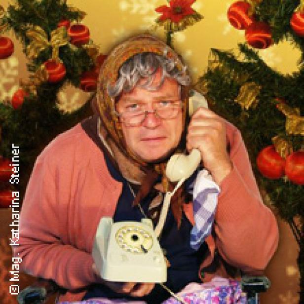 Eine ältere Person in Frauenkleidung telefoniert vor einem Weihnachtsbaum.