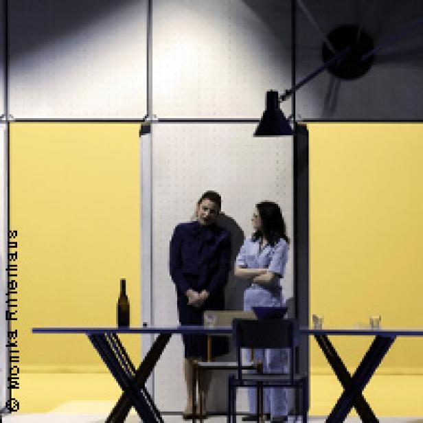 Zwei Frauen stehen in einem Raum mit gelben Wänden und sprechen miteinander.