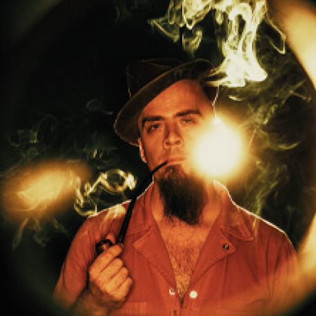 Ein Mann mit Bart und Hut raucht eine Pfeife, umgeben von Rauchschwaden.