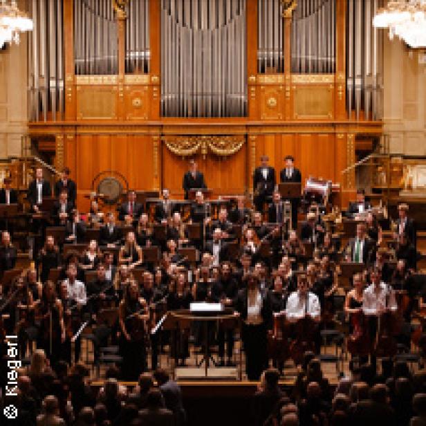 Ein Orchester spielt vor einem Publikum in einem Konzertsaal mit einer großen Orgel im Hintergrund.