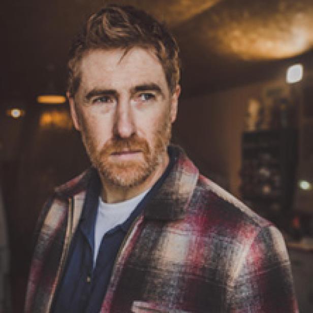 Ein Mann mit rötlichem Haar und Bart trägt ein kariertes Hemd.