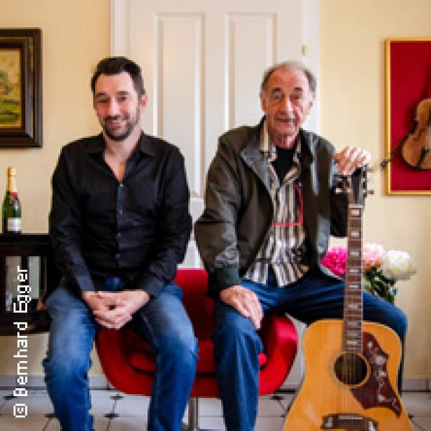 Zwei Männer sitzen vor einer Tür, einer hält eine Gitarre.