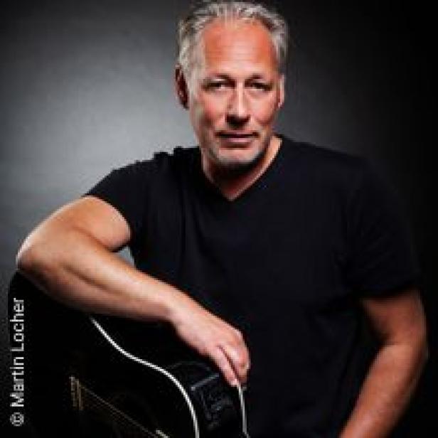 Ein Mann mit grauen Haaren und schwarzem T-Shirt posiert mit einer Gitarre.