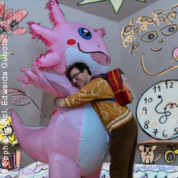 Ein Mann umarmt einen großen, aufblasbaren, rosa Axolotl in einem Zimmer mit Wandmalereien.