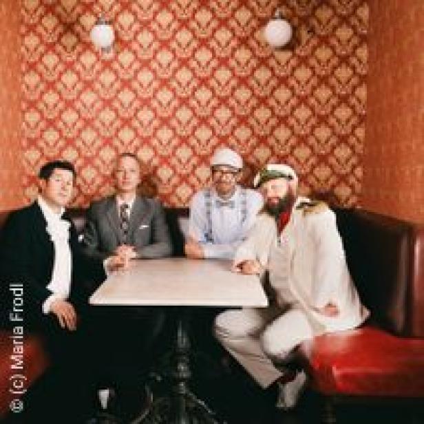 Vier Männer sitzen an einem Tisch in einem Raum mit gemusterter Tapete.