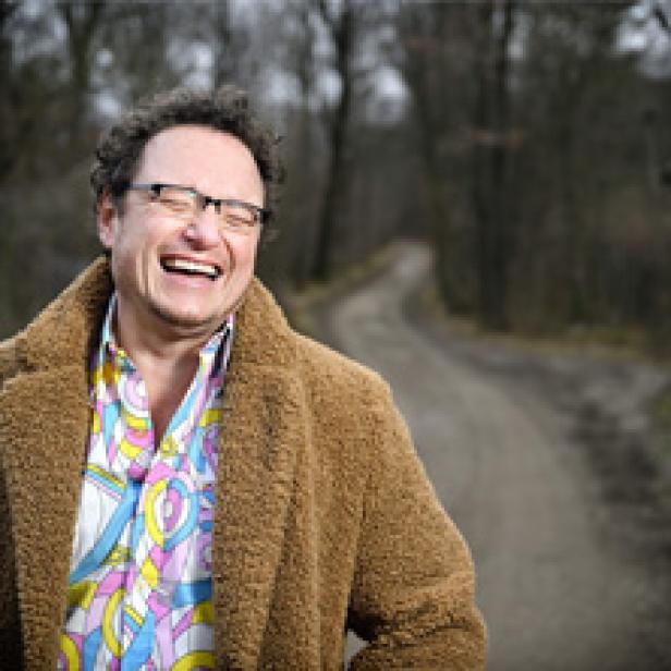 Ein lachender Mann mit Brille und braunem Mantel steht auf einem Waldweg.