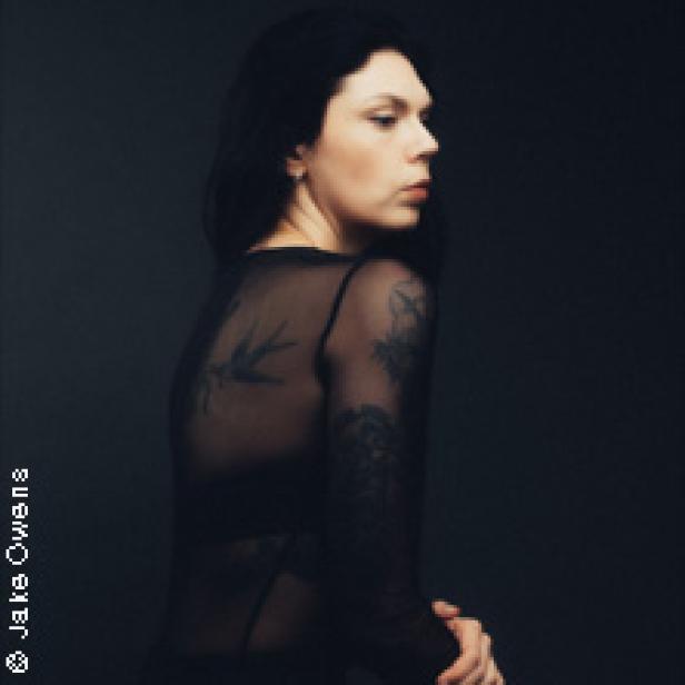 Eine Frau mit dunklen Haaren und Tätowierungen blickt über ihre Schulter.