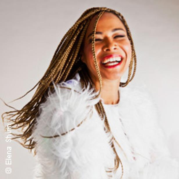 Eine lachende Frau mit geflochtenen Zöpfen und einem weißen, federartigen Oberteil.