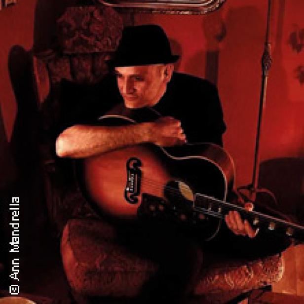 Ein Mann mit Hut sitzt in einem Sessel und hält eine Gitarre.