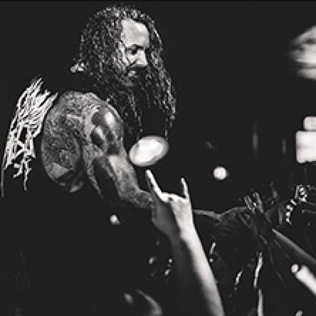 Ein Mann mit langen Haaren und Tätowierungen macht auf einer Bühne die Pommesgabel.