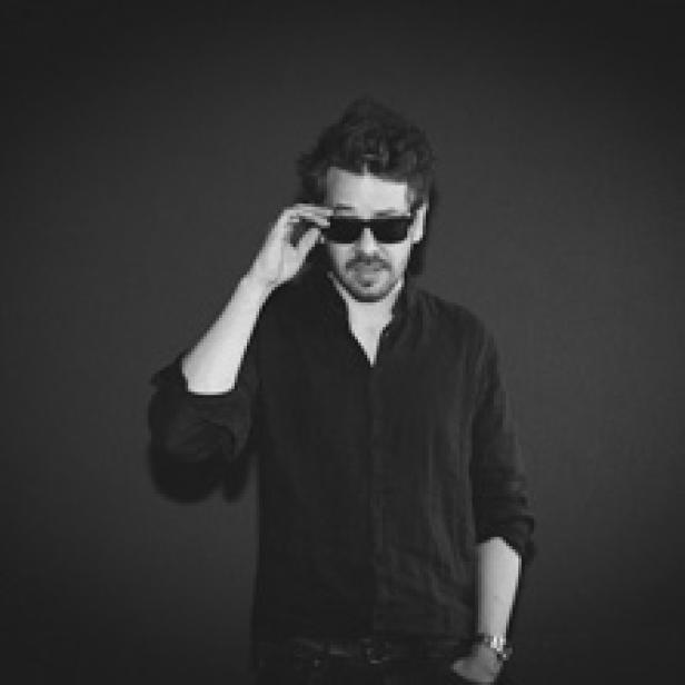 Ein Mann mit Sonnenbrille posiert vor einem dunklen Hintergrund.