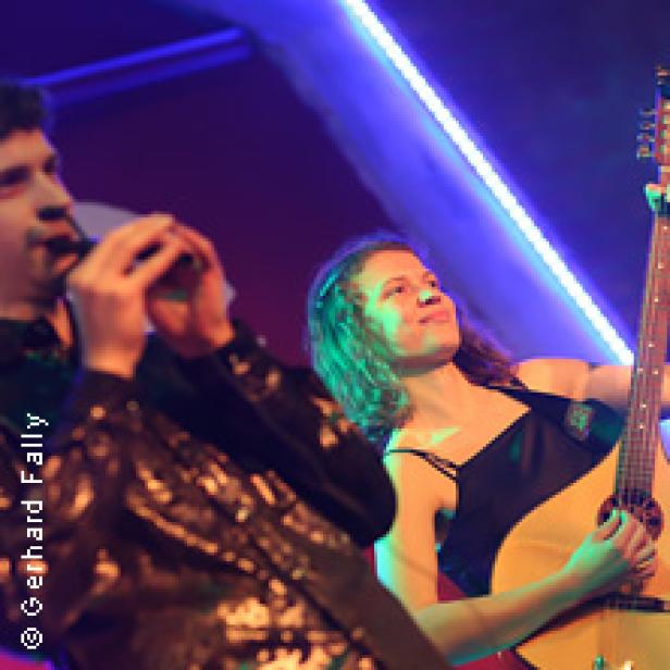 Eine Frau mit Gitarre und ein Mann mit einem Blasinstrument treten auf einer Bühne auf.