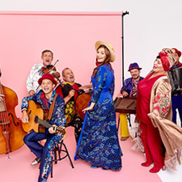 Eine Gruppe von Musikern in farbenfrohen Kostümen spielt auf verschiedenen Instrumenten vor einem rosa Hintergrund.