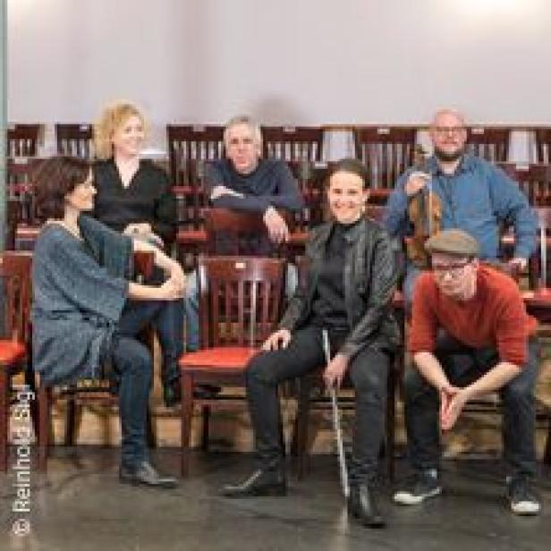 Eine Gruppe von sechs Personen sitzt und steht in einem Raum mit Stuhlreihen.