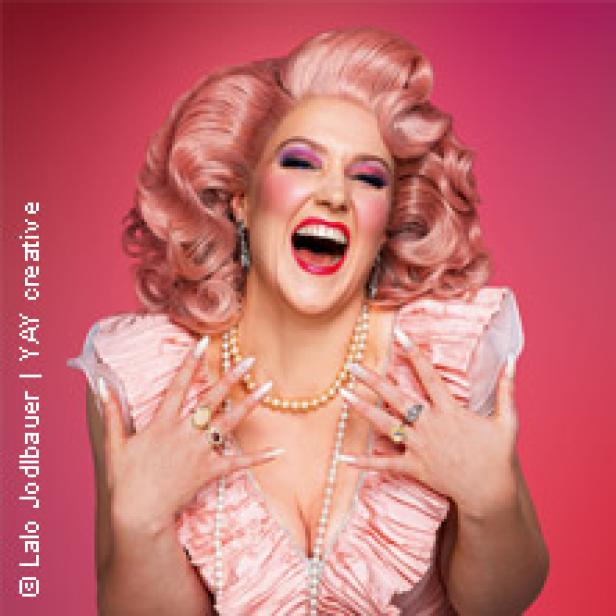Eine Dragqueen mit rosa Perücke, üppigem Make-up und einem rosa Kleid lacht.