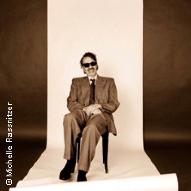 Ein Mann mit Sonnenbrille sitzt in einem Anzug auf einem Stuhl vor einem neutralen Hintergrund.