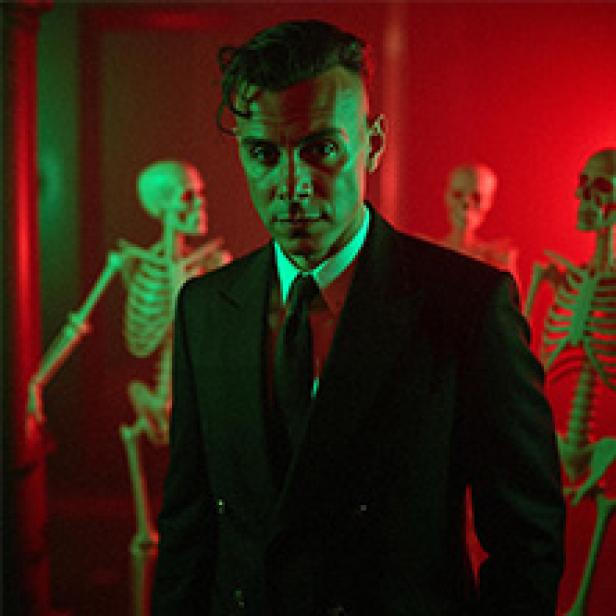 Ein Mann in einem Anzug steht vor drei Skeletten, beleuchtet in Rot und Grün.