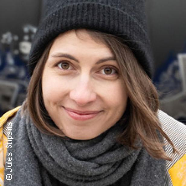 Eine Frau mit braunen Haaren, Mütze und Schal lächelt in die Kamera.