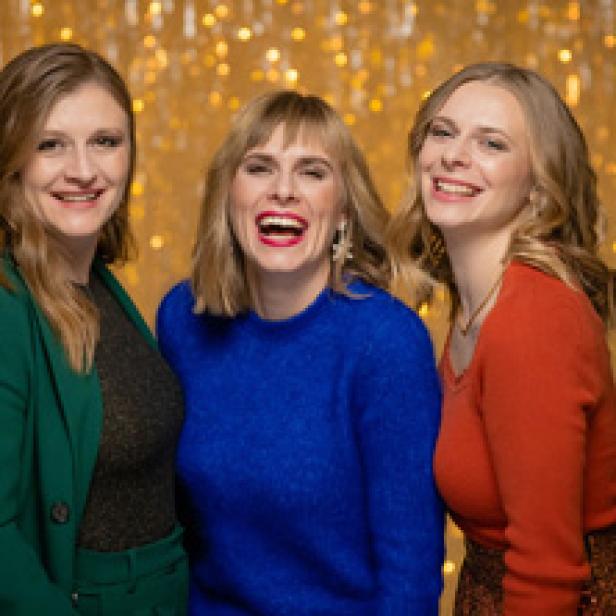 Drei Frauen stehen vor einem goldenen Hintergrund und lachen.