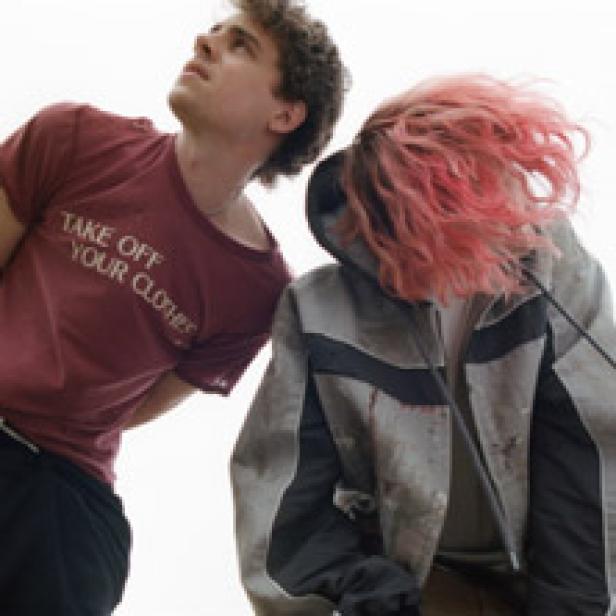 Ein junger Mann mit einem roten T-Shirt steht neben einer Person mit pinkfarbenen Haaren.