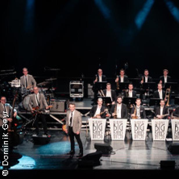 Eine Big Band tritt auf einer Bühne vor dunklem Hintergrund auf.