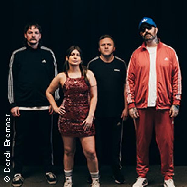 Eine Band posiert vor einem schwarzen Hintergrund, alle tragen Trainingsanzüge oder sportliche Kleidung.