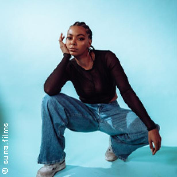 Eine junge Frau in Jeans und einem schwarzen Oberteil posiert vor einem hellblauen Hintergrund.