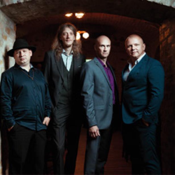 Vier Männer in Anzügen stehen in einem gewölbeartigen Raum.