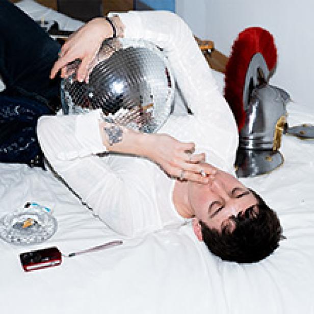 Ein Mann liegt mit einer Discokugel und einem römischen Helm auf einem Bett und raucht.