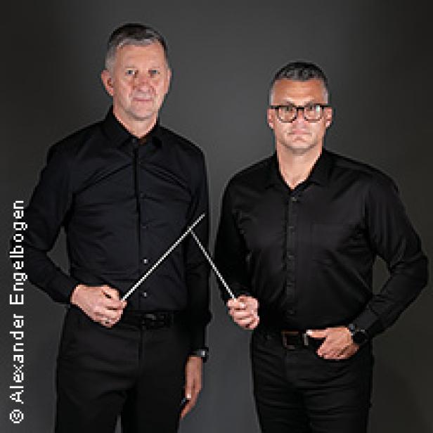 Zwei Männer in schwarzen Hemden halten Dirigentenstäbe vor einem grauen Hintergrund.