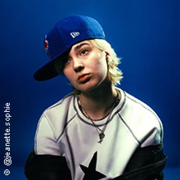 Eine junge Frau mit kurzer, blonder Haare und einer blauen Baseballkappe posiert vor einem blauen Hintergrund.