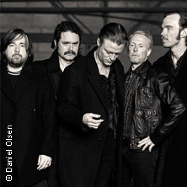 Die Band Interpol steht in dunkler Kleidung vor einer grauen Wand.
