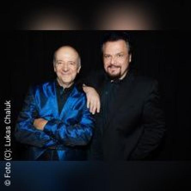 Zwei Männer, einer mit blauem Sakko, posieren vor einem schwarzen Hintergrund.