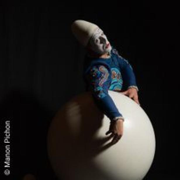 Ein Clown mit weißem Gesicht und blauem Kostüm sitzt auf einer großen, weißen Kugel.