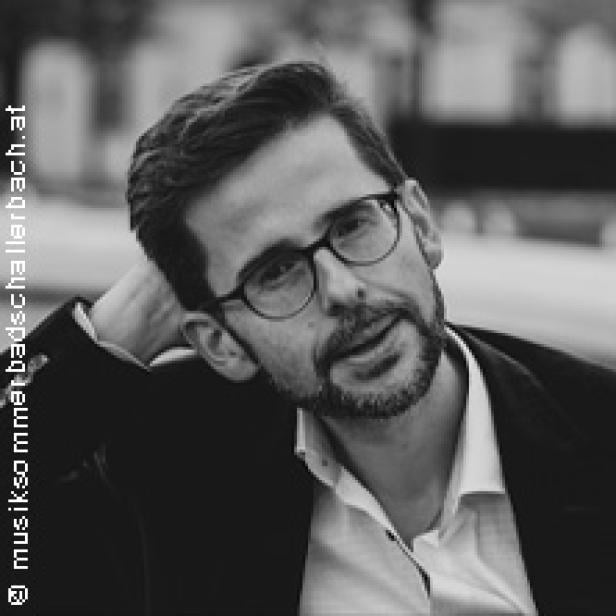 Ein Mann mit Brille und Bart, in Jackett und Hemd, lehnt entspannt.