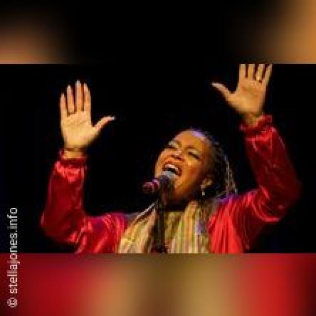 Stella Jones singt mit erhobenen Händen in ein Mikrofon vor einem dunklen Hintergrund.