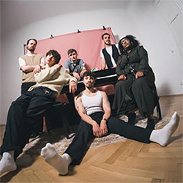 Eine sechsköpfige Band sitzt und posiert vor einem rosa Hintergrund.