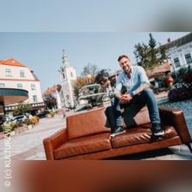 Ein Mann sitzt auf einer braunen Couch vor einem Platz mit Kirche.
