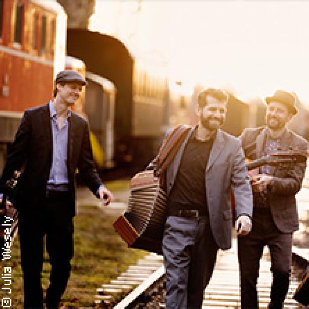 Drei Männer in Anzügen gehen mit Musikinstrumenten an Bahngleisen entlang.